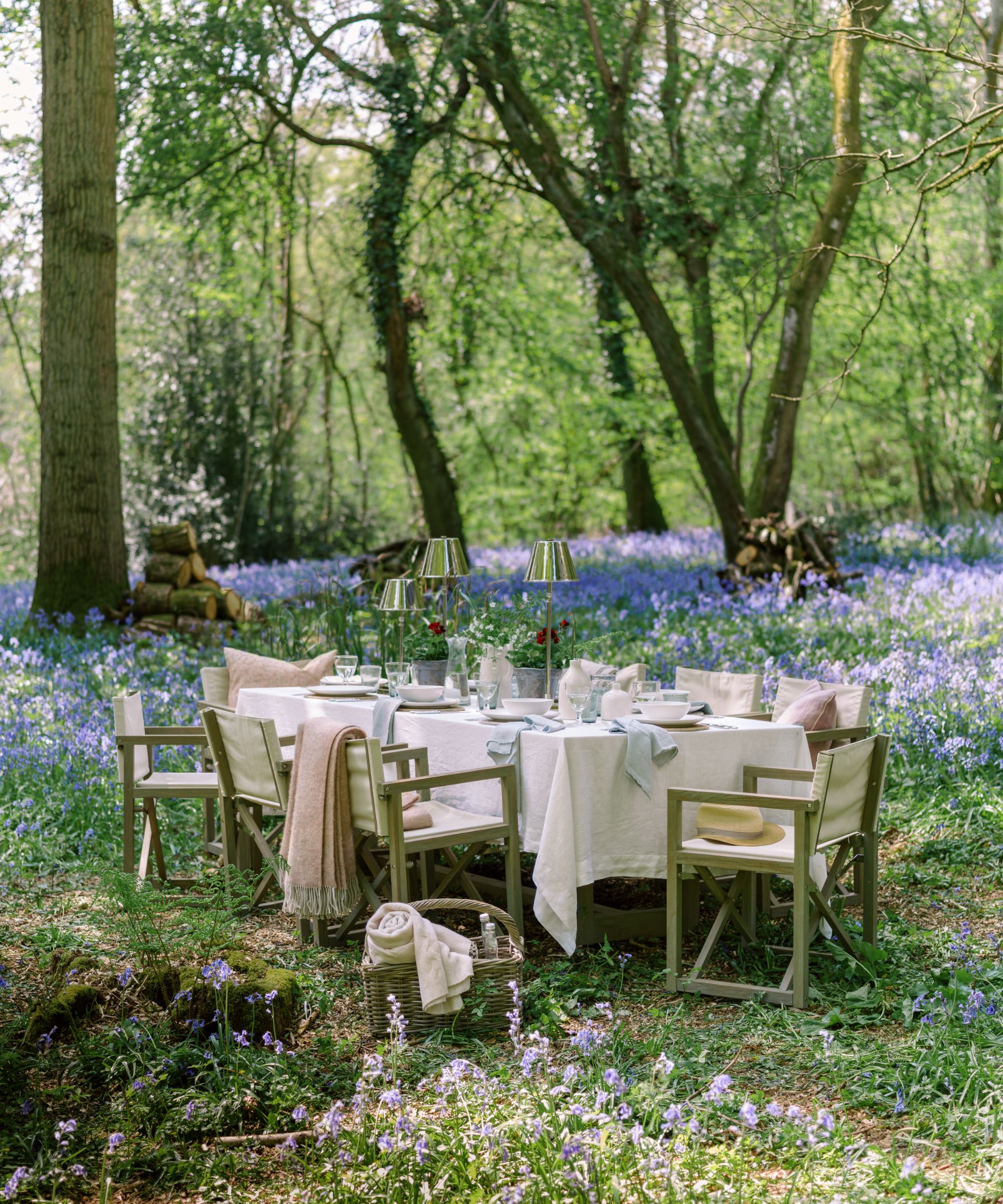 large outdoor table set up in a woodland with bluebells the table is laid with white linens and silver portable table lamps