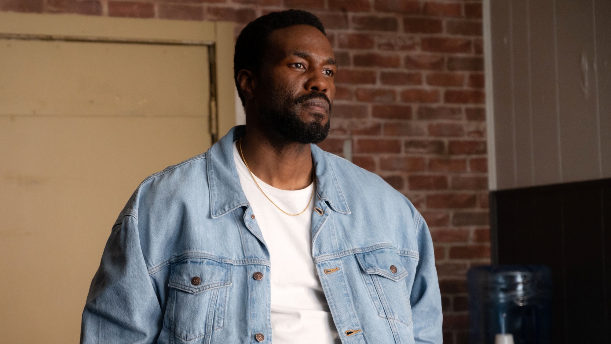 Yahya Abdul-Mateen II as Simon looking serious in a white t-shirt and light blue denim jacket in Wonder Man.