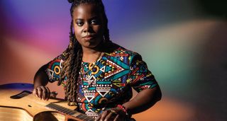 Yasmin Williams plays her acoustic guitar on her lap, and wears a brightly patterned shirt, against a brightly textured color background.