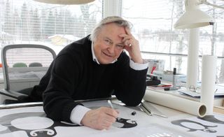 Eero Aarnio loking into the camera and smiling, while sketching at his desk