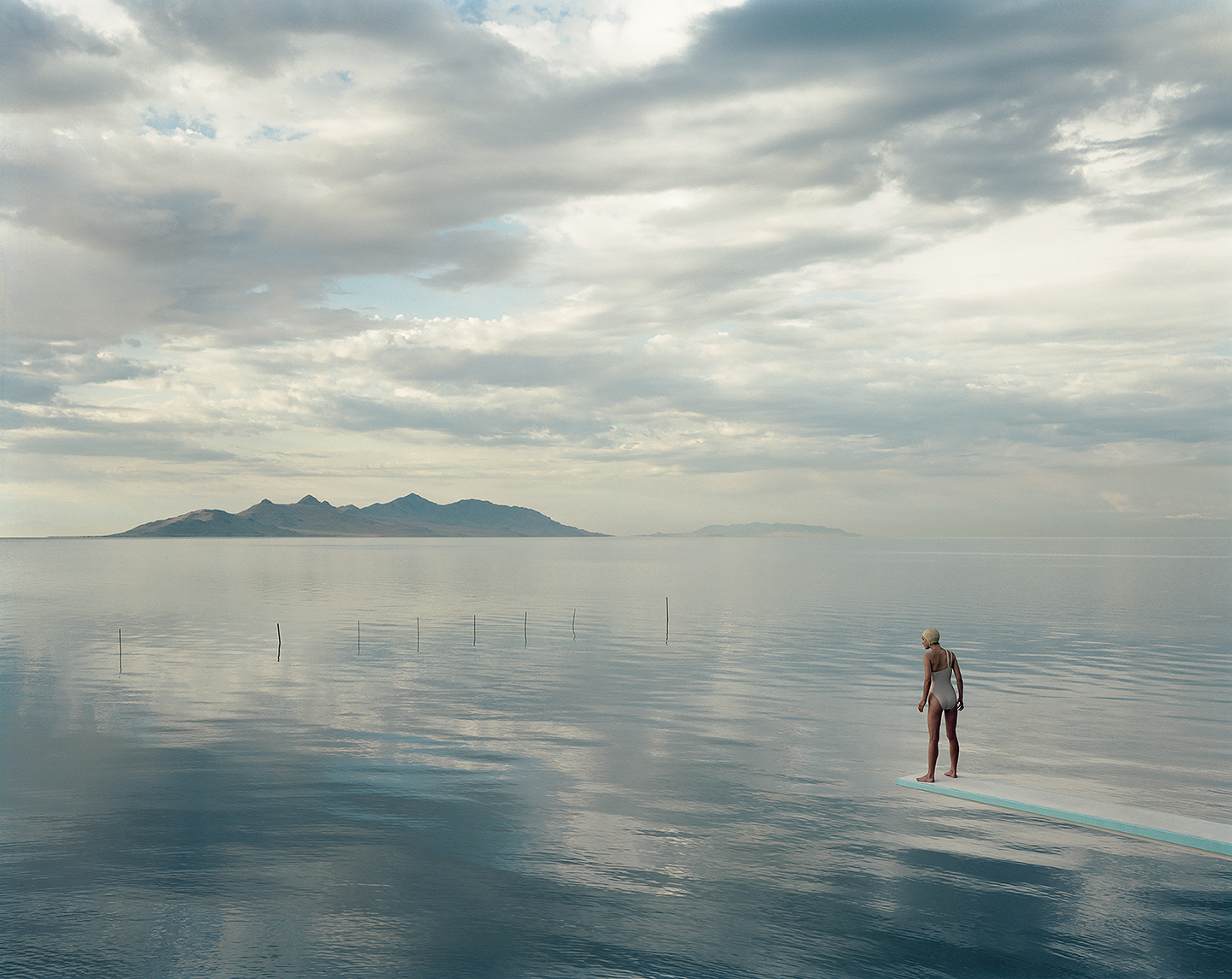 A woman in a swimsuit and cap stands at the end of a diving board that seems to float above a vast, still lake reflecting a cloudy sky, with mountains on the distant horizon.