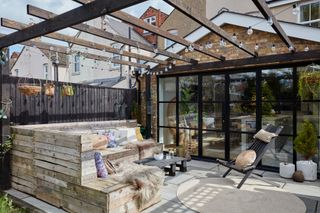 Rear extension with Crittall-style doors leading to patio area with black painted pergola, pallet L-shaped seating and festoon lights hanging above