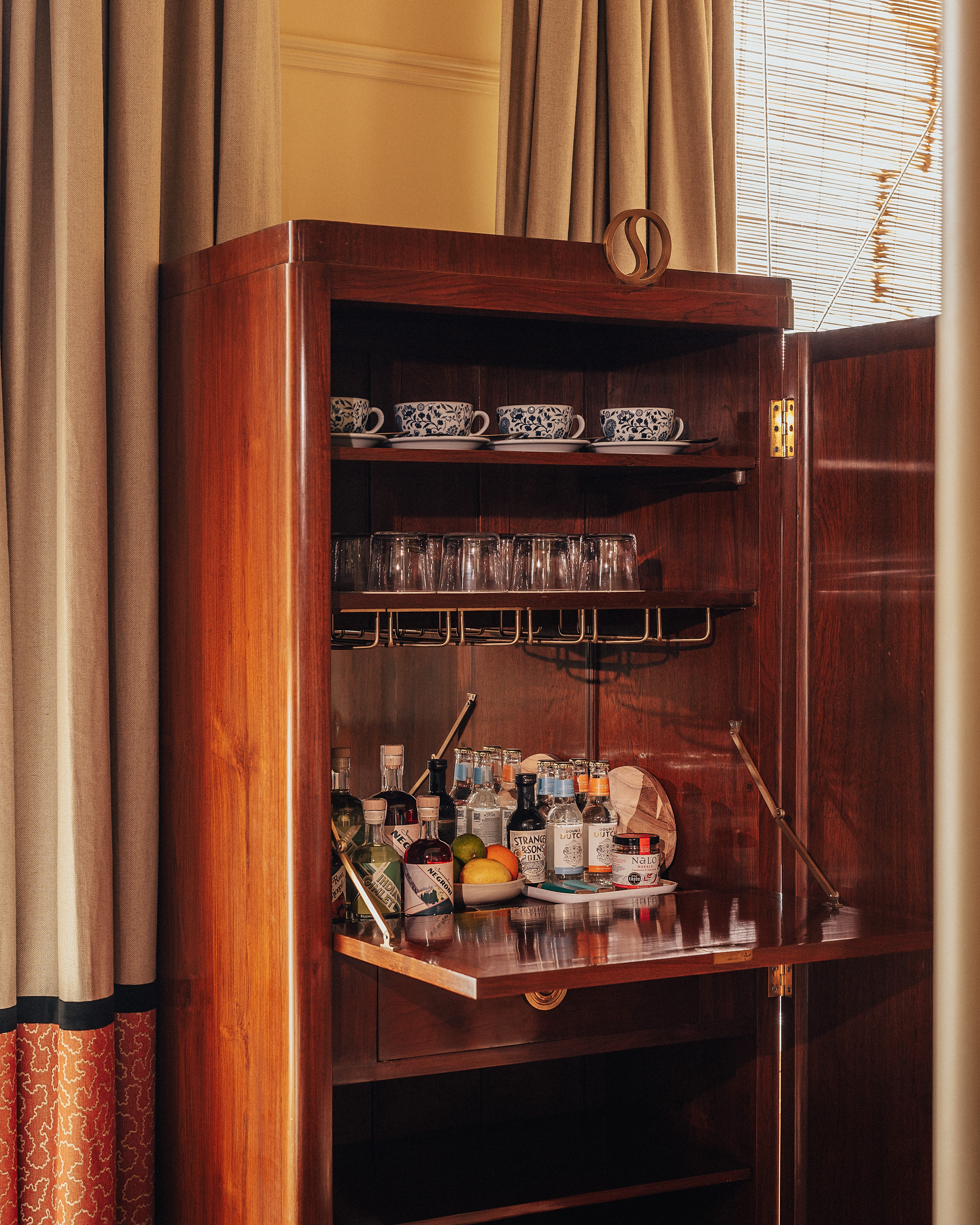 A layered, transitional hotel suite featuring a series of figurative, colorful artworks on its walls, patterned furniture, including rugs, armchairs, and sofas, split-color pillars, and a buttery, tile blue, and green palette.