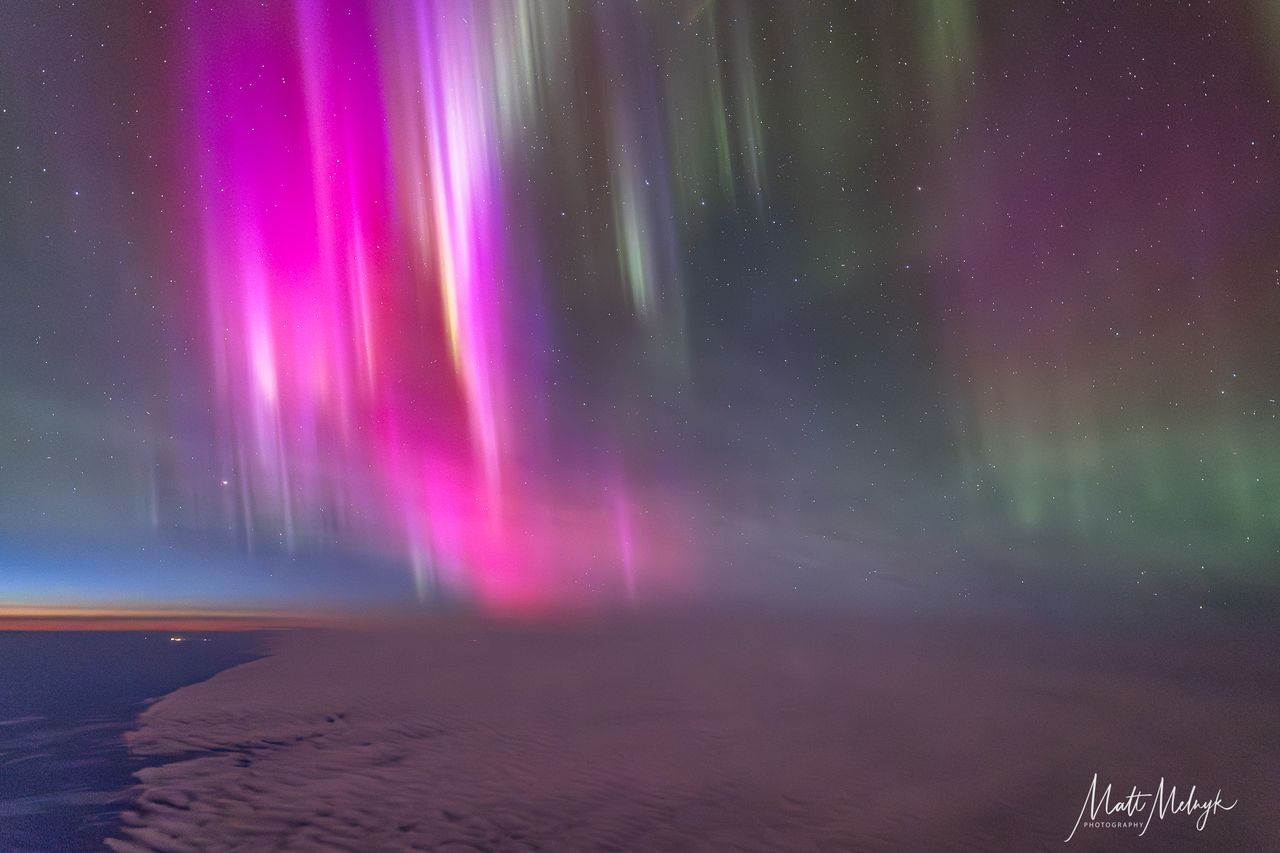 curtains of magenta and green light dance in the sky, a thin blue line marking Earth's atmosphere is visible in the distance.
