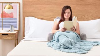 Image shows a woman with long brown hair reading a book while leaning back on a Husband pillow in bed