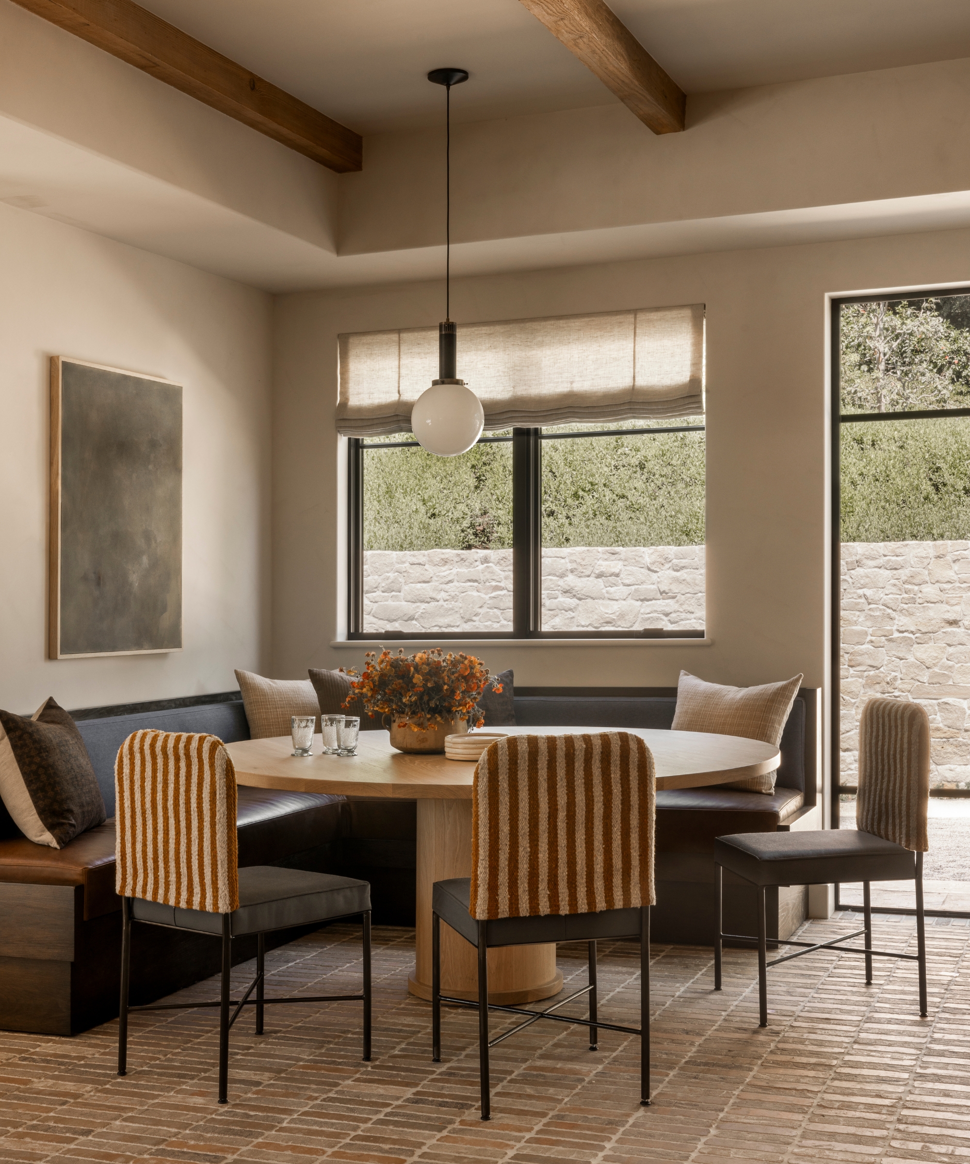 A kitchen nook with charcoal L-shaped banquette seating, a round wood pedestal table, stripe back chairs, and a white globe pendant above