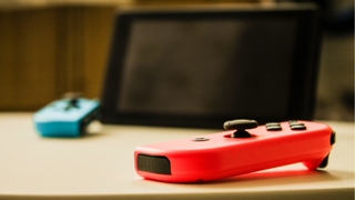 Un Joy-Con rojo neón sobre una mesa en primer plano, con una Nintendo Switch y un Joy-Con azul de fondo.