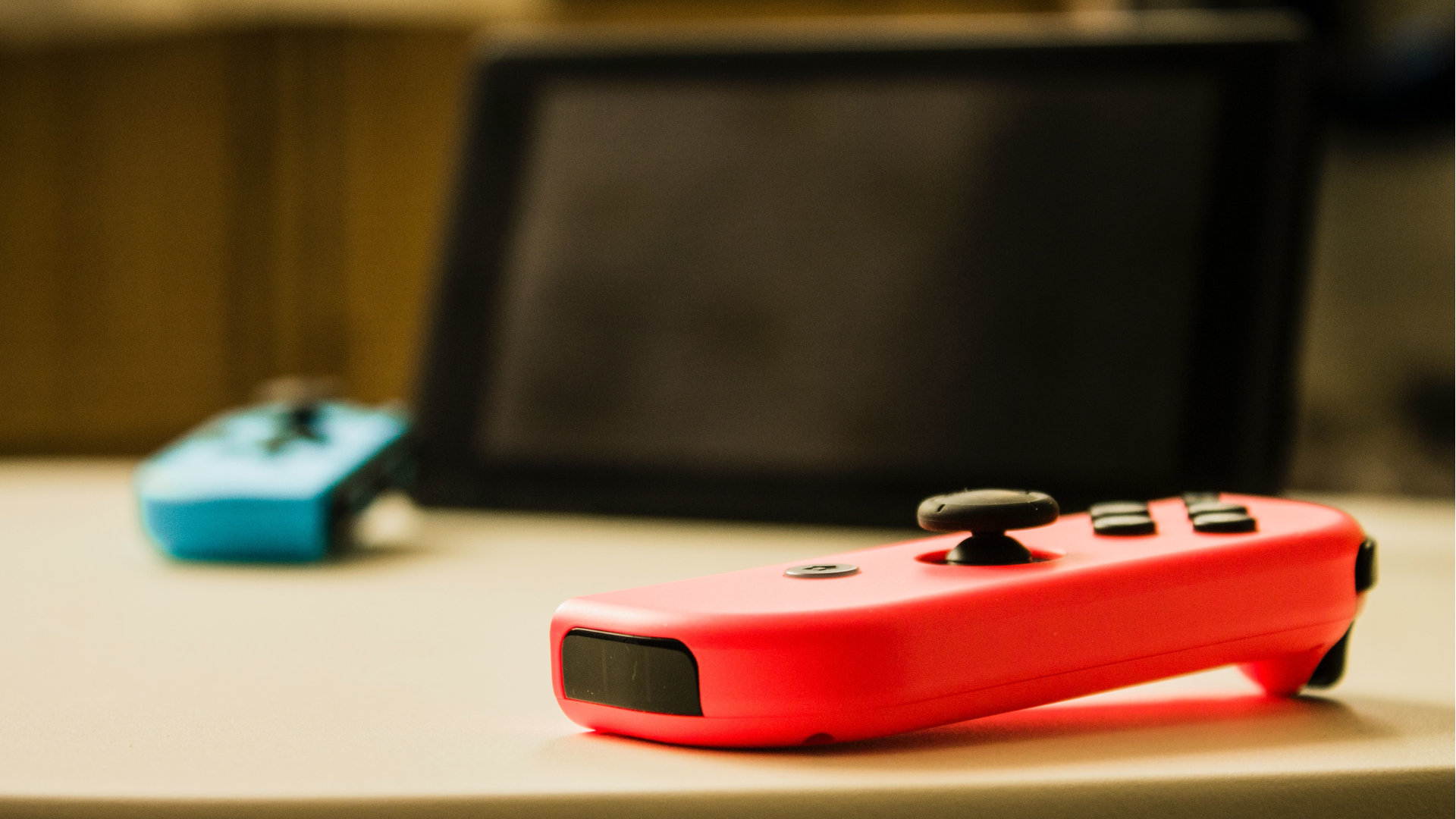 Неоново-красный Joy-Con на столе на переднем плане, с экраном Nintendo Switch и синим Joy-Con, лежащим на столе на заднем плане, немного не в фокусе