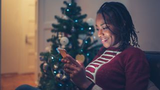 Woman looking at mobile phone, sitting on couch in the living room with glittering Christmas tree behind her