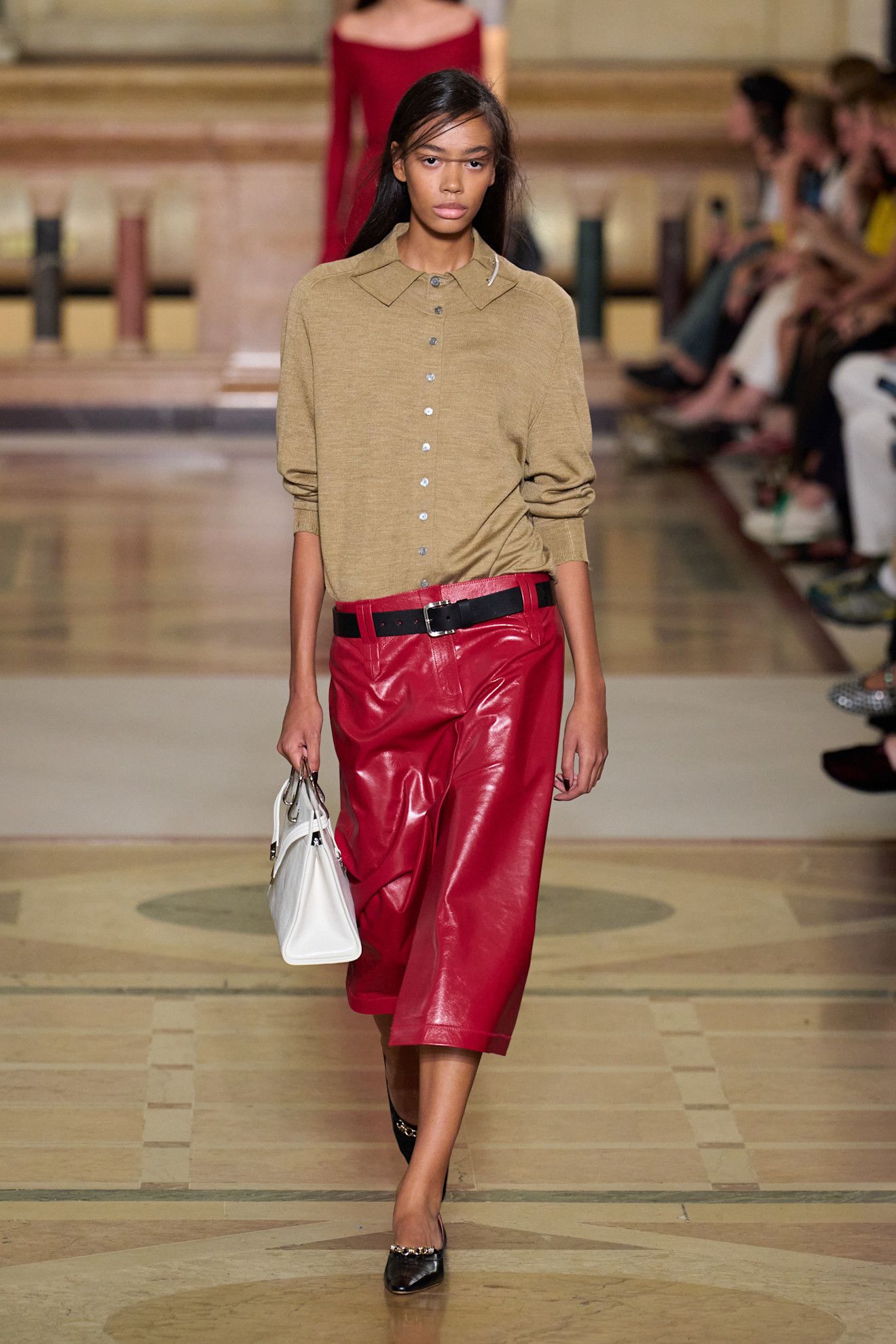 A model wears a beige cardigan, black belt, red leather skirt, white bag, and black heels.