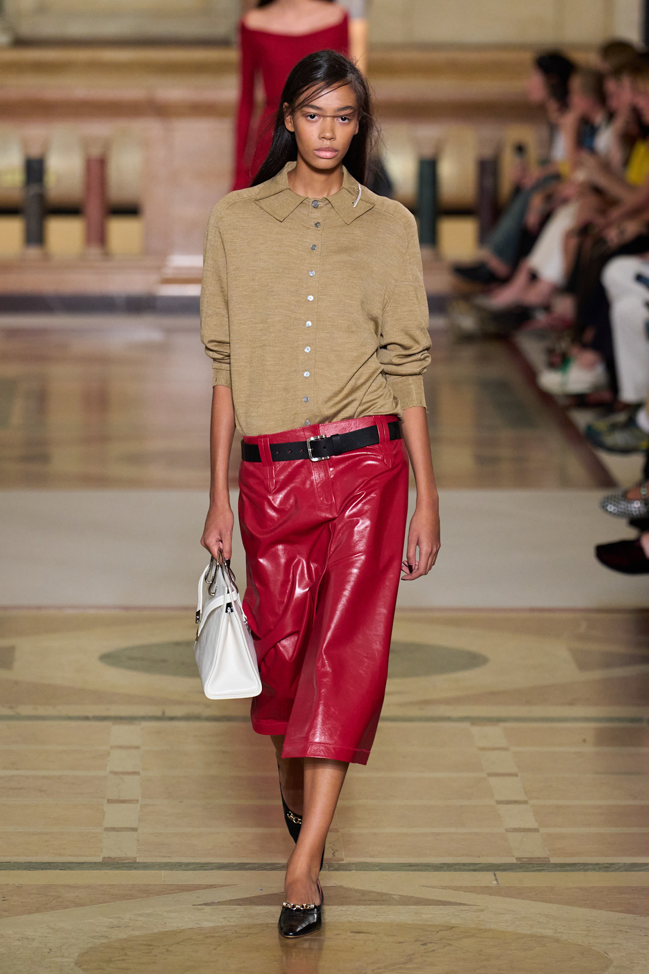 A model wears a beige cardigan, black belt, red leather skirt, white bag, and black heels.