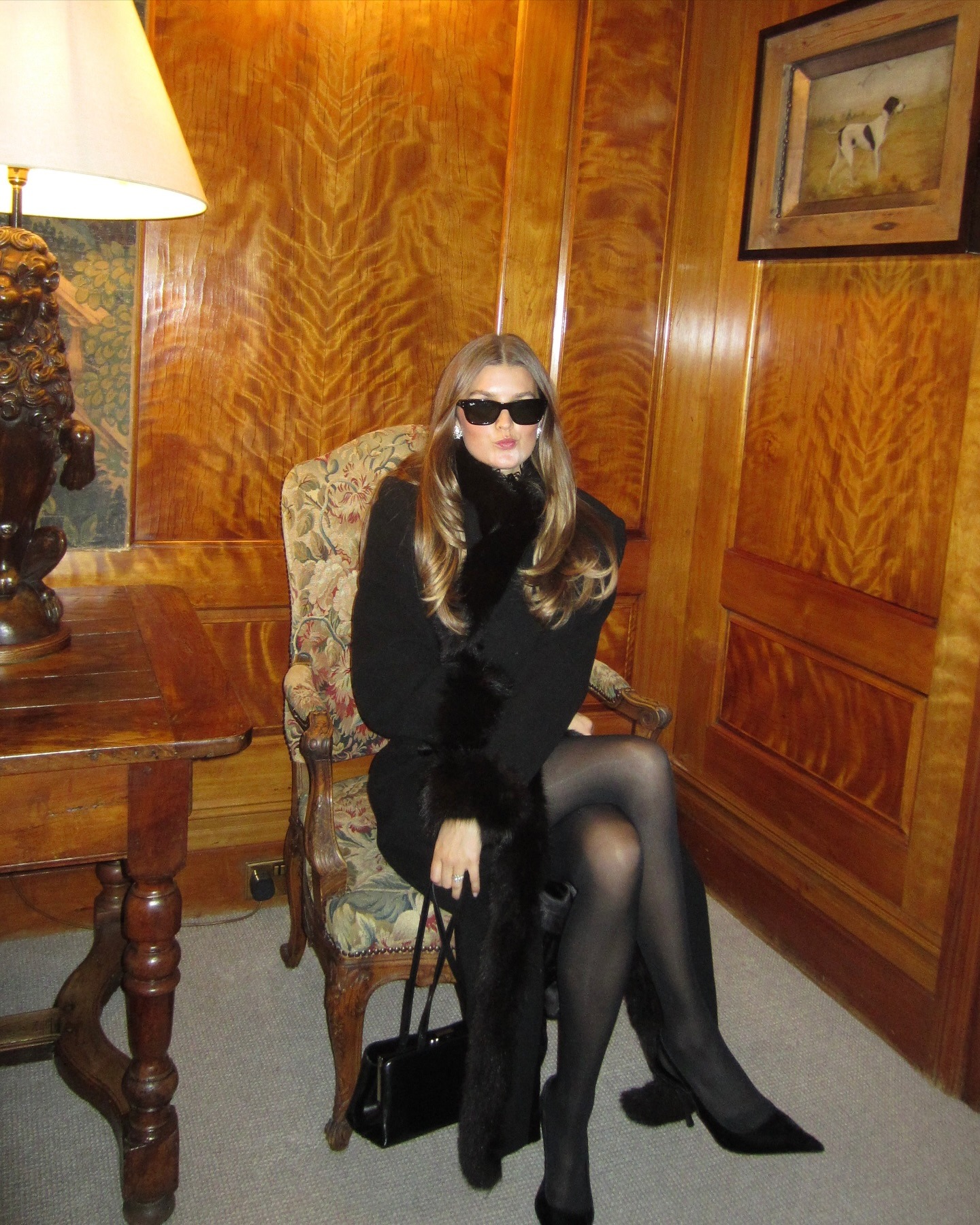 Sara wears sheer tights, pumps, and a black coat with fur trim.