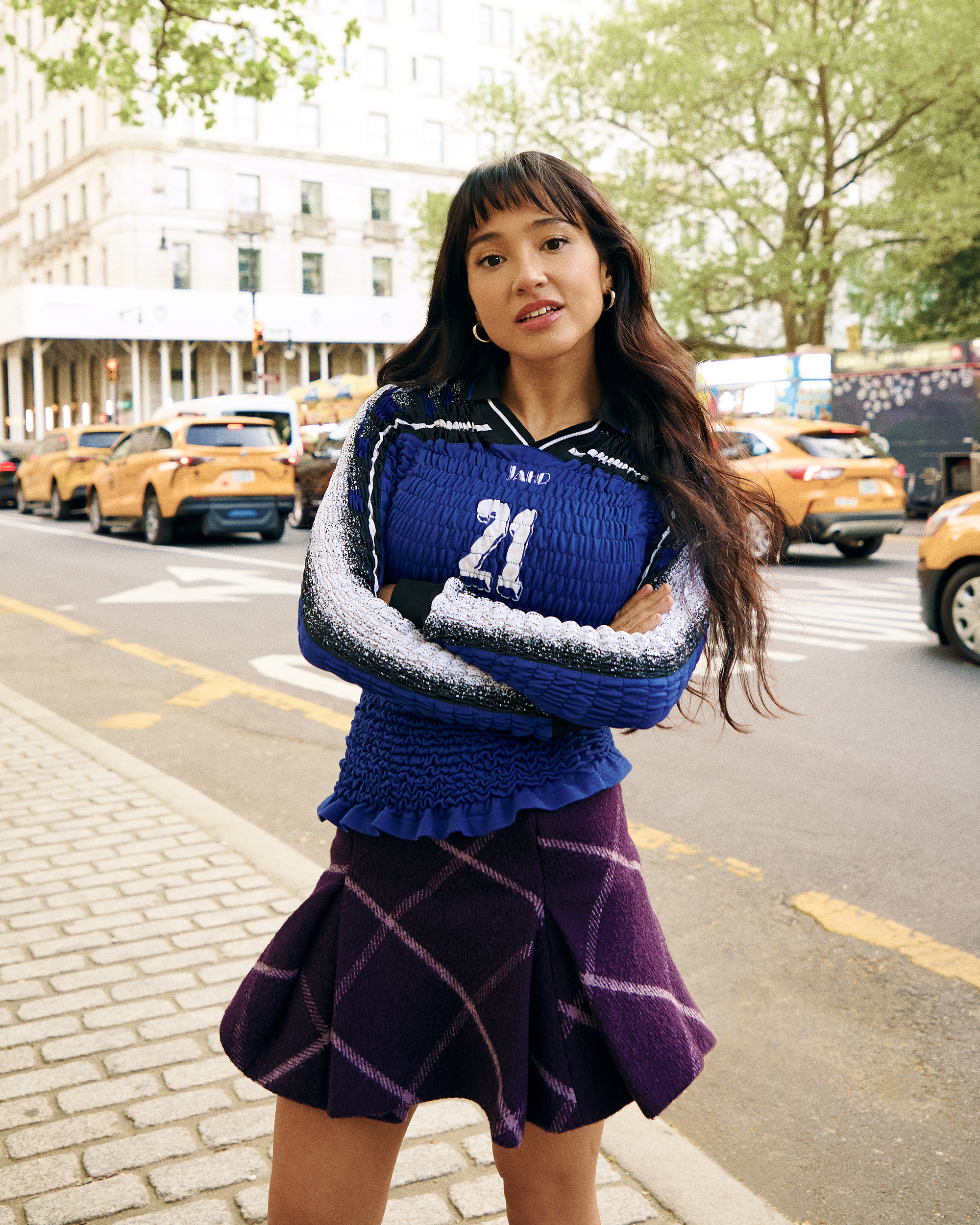A atriz Wally Baram, supercompensadora, posa para um retrato no Plaza Hotel, em Nova York. Ela veste uma camisa azul de manga comprida com o número 21 na frente e uma minissaia xadrez roxa com botas de couro pretas que vão logo abaixo do joelho.