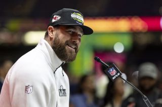 Travis Kelce wearing a white zip fleece and black Kansas City Chiefs Super Bowl hat talking into a microphone and smiling at a press conference