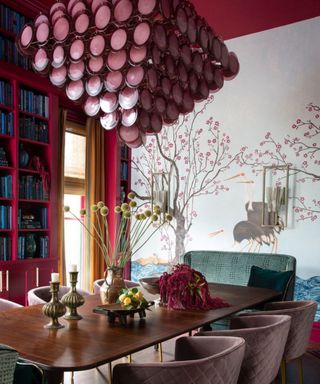 A dining room with tall burgundy bookshelves, a dark wooden table with gold candles and a vase of flowers on it, six purple chairs around it, and a wall mural with cherry blossoms and a heron bird
