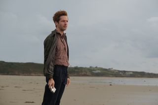 Jim Bergerac (Damien Molony) stands on the beach - it's clearly windy because his hair is being blown back and the front of his jacket, which is unbuttoned, has been blown back against his arms. In his right arm he is holding a silver hip flask, upside down.