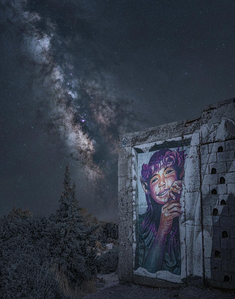 A mural of the Greek myth Pandora painted on a stone slab sits in the foreground of an image of the milk way, shooting up diagonally from the murals corner.