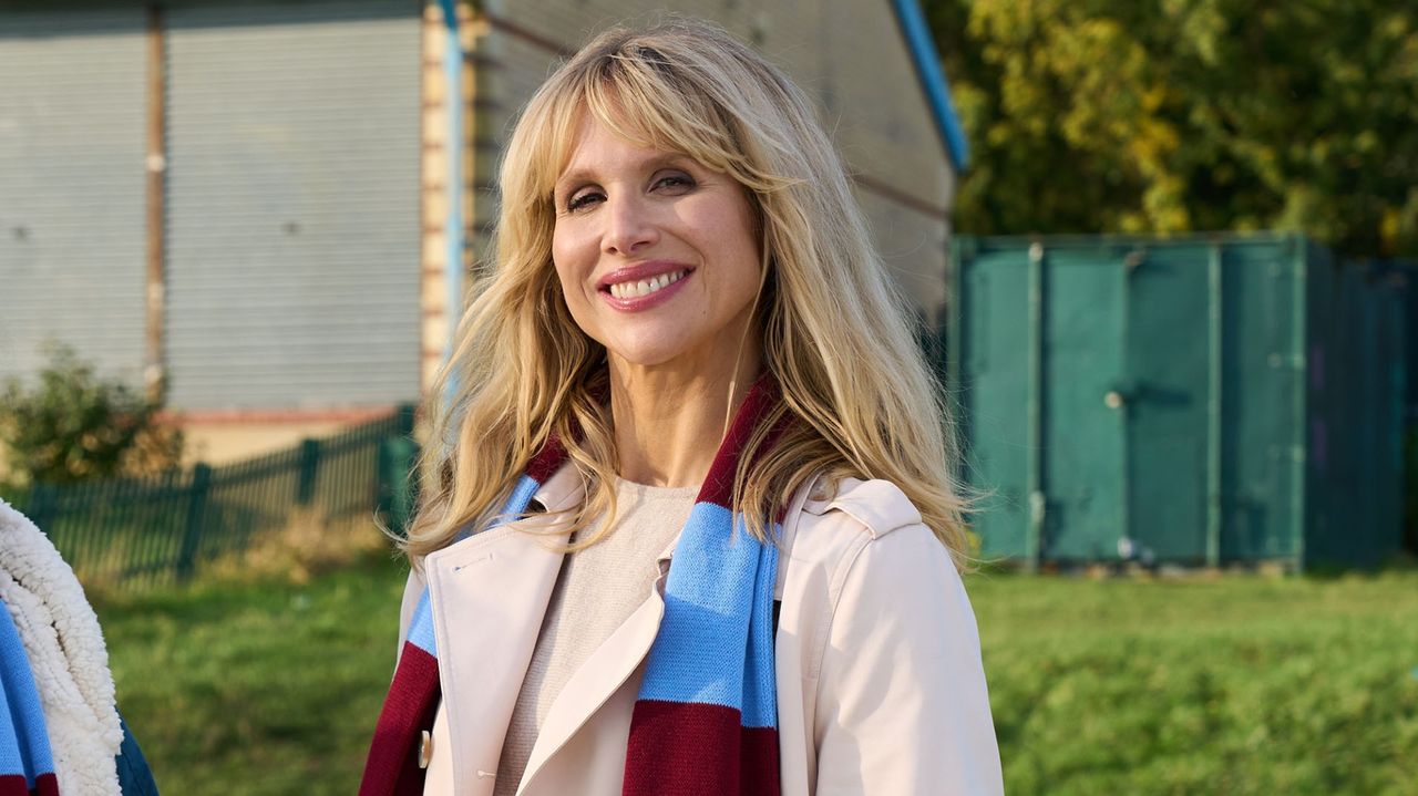 Amanda smiles, wearing a trench coat and football scarf, in Amandaland