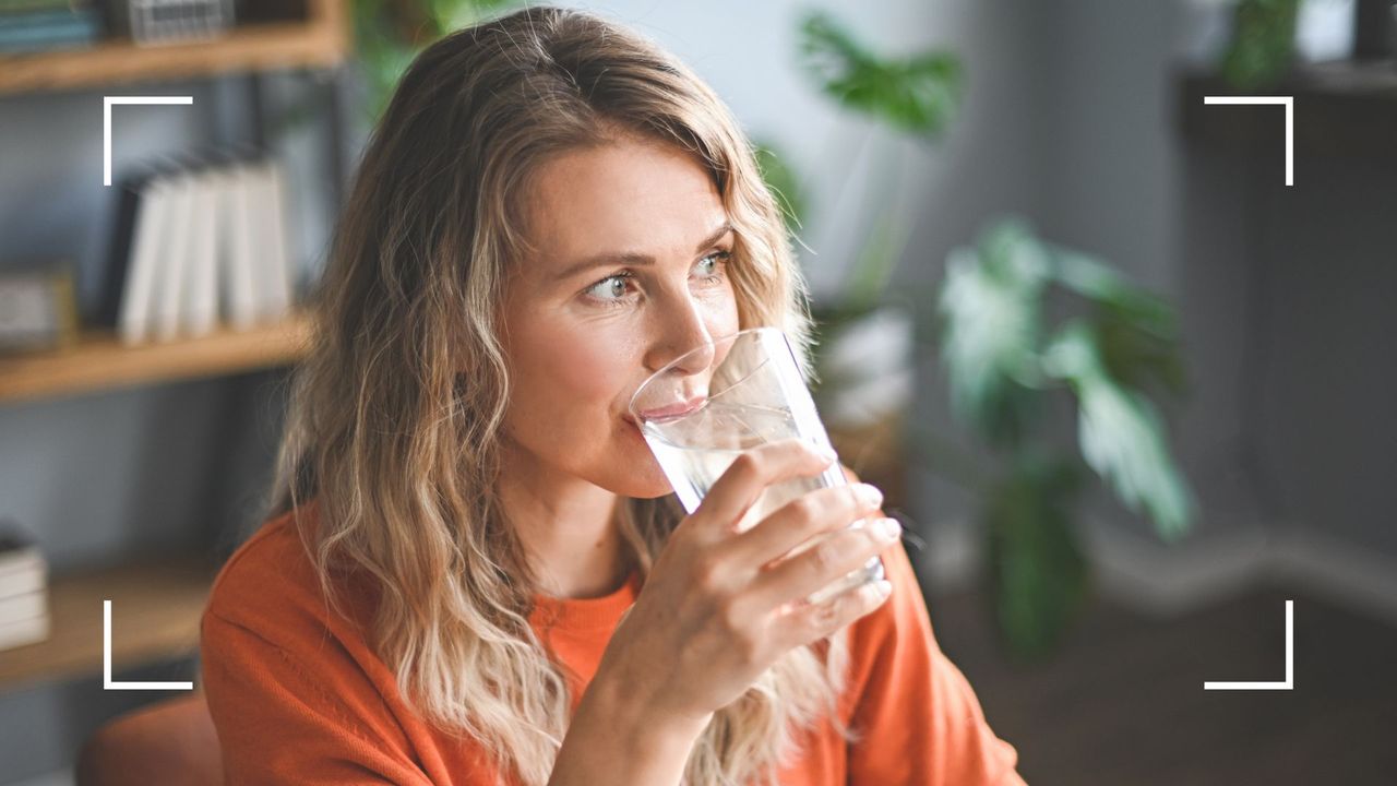 Woman sipping from a glass of ice water, sitting at desk with a bookshelf and plants behind her, representing the link between alcohol and menopause