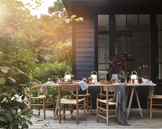 Outdoor dining ideas with wooden table and chairs outside black wood panelled house, with autumnal table decor and grey blankets