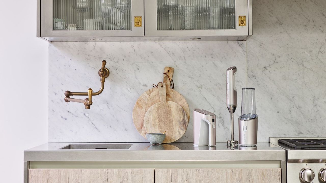 A white blender set with single cup blender and a stick blender sitting on a kitchen countertop between a sink with a pot filler arm and a stove, a marble backsplash and two leaning cutting boards behind them. Glass flued cabinets above. 