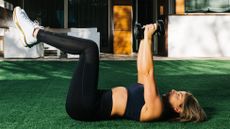 A woman lies outside on fake grass wearing sports clothes, with her hands in the air clutching a pair of dumbbells. Her legs are bent at 90° so her shins are parallel to the floor. 