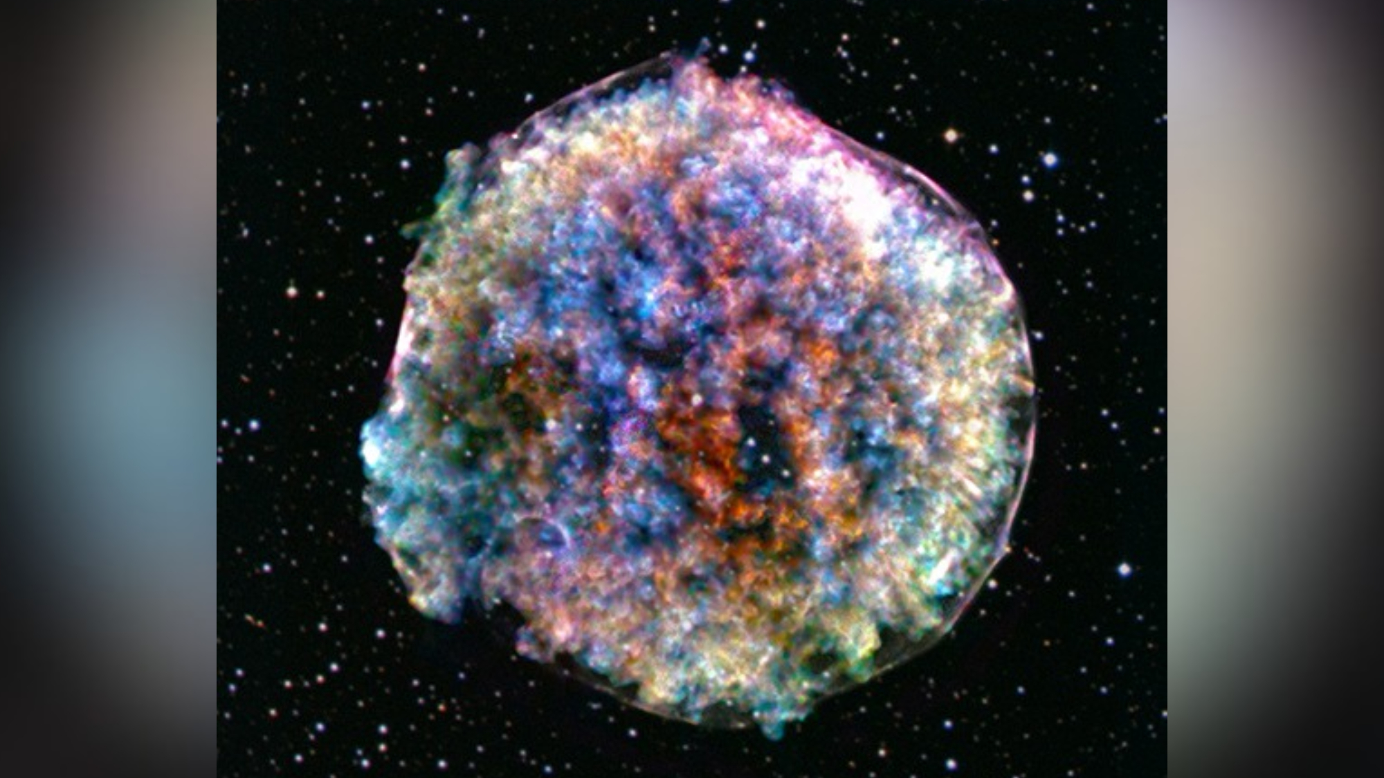 A circular cloud of glowing red, orange and blue stellar debris against a black, star-filled background, with a bright blue outer rim and colorful clumps scattered throughout.