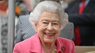 Here we see Queen Elizabeth II with a big smile on her dace, causing her eyes to wrinkle up. She has short snow white hair and is wearing a pearl necklace, floral dress, and a pink blazer with a gold and purple floral brooch.