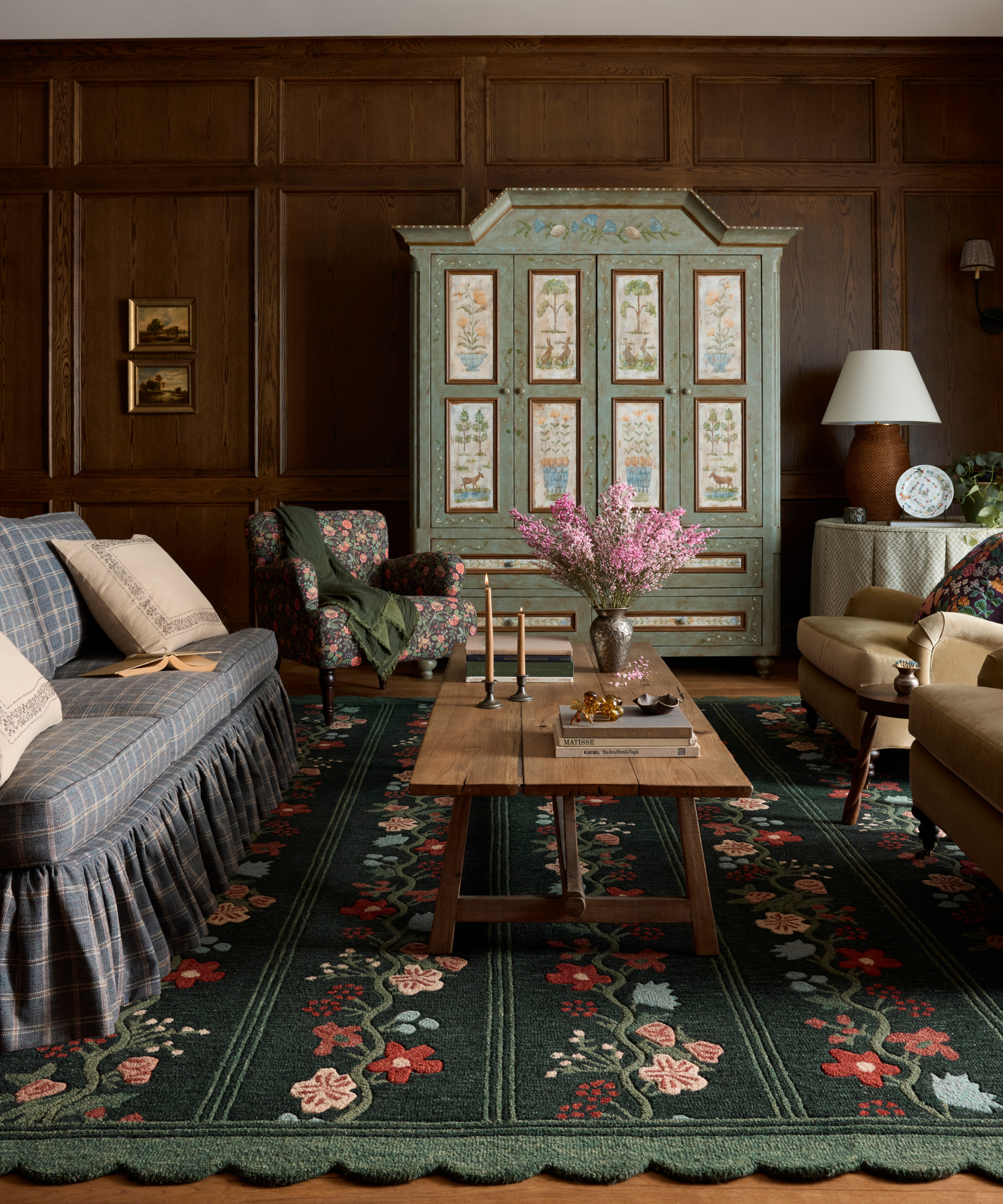 a living room with wood paneled walls, a large painted cabinet, a wooden coffee table, a floral rug, and a plaid couch