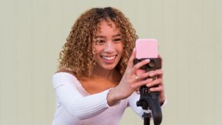 female blogger with curly hair recording video for vlog on modern cellphone while standing on green background on street