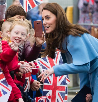 Kate Middleton wearing a blue cape coat leaning forward to talk to kids waving Union Jack flags