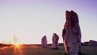 the sun rises in the distance, illuminating three large oblong stones standing on end in the grass