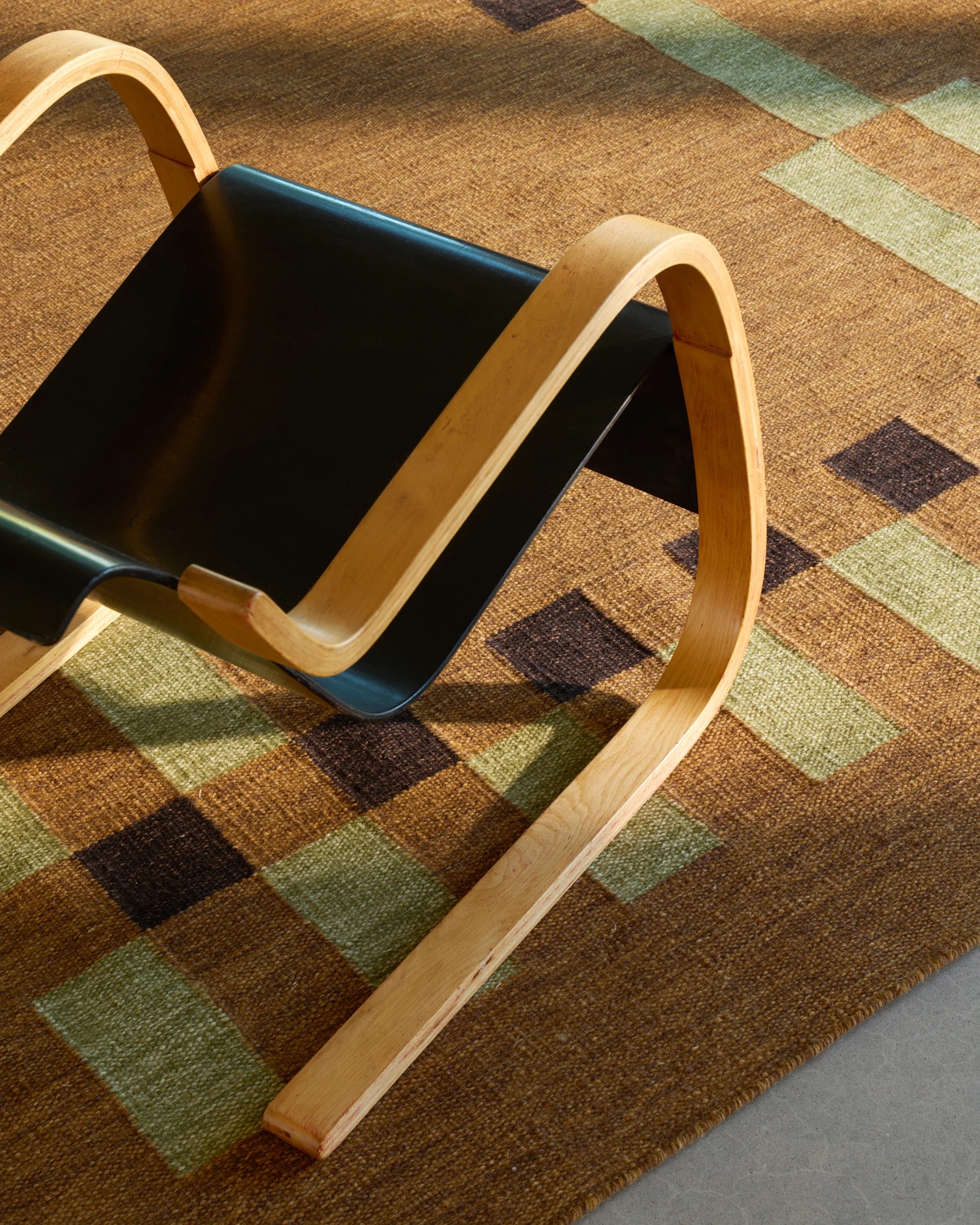 Tradition 02 Runner rug in brown, green, and ochre in a sun-lit living room, topped by a black and pale wood chair.