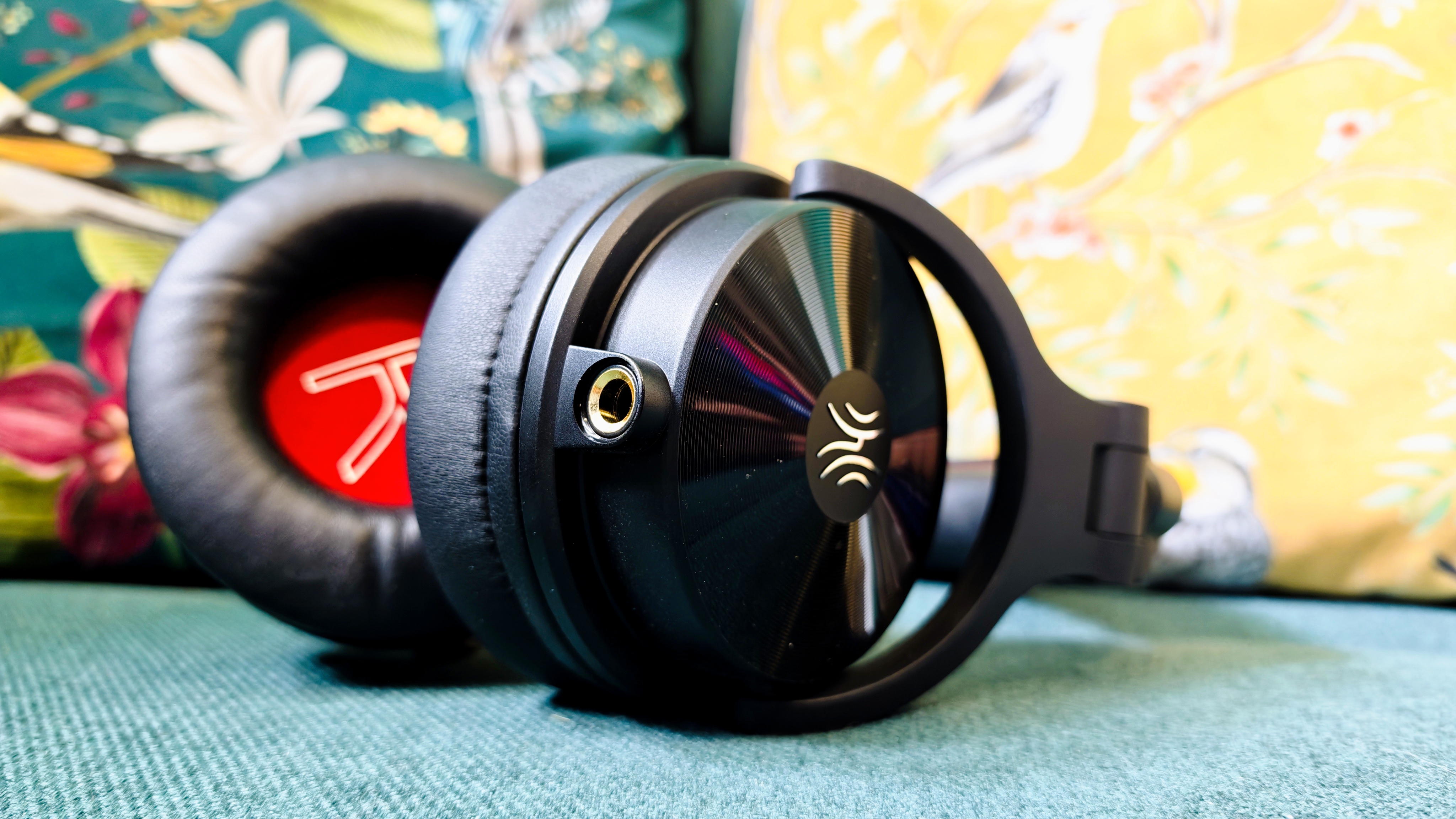 A pair of black OneOdio Studio Max 2 headphones with red internal earcups sitting on a green sofa in front of colourful pillows with birds and flowers on them