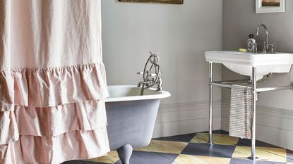 A bathtub with a pink frilly shower curtains wrapped around it, grey painted walls, a standing sink, and a blue and yellow tiled checker floor