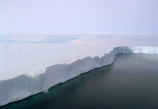 An in-flight photo shows the edge of the Larsen B Ice Shelf. Pools of meltwater can be seen on the surface, and ice flows off the edge like a waterfall.
