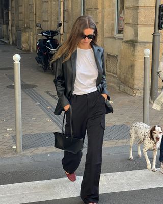 French influencer walking on street wearing white t-shirt, black pants, and black leather blazer.