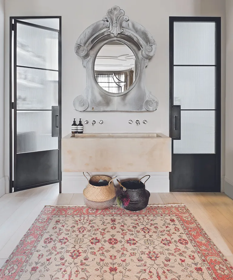 Master en suite with oriental rug with floral pattern on wooden floor, oval 19th century French mirror in ornate grey frame over large stone basin framed by Crittal doors.