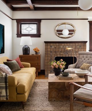 Cozy living room with dark wooden window frames, brick fireplace, yellow sofa, wooden coffee table, armchair, cozy cushions, blankets and decorative ornaments