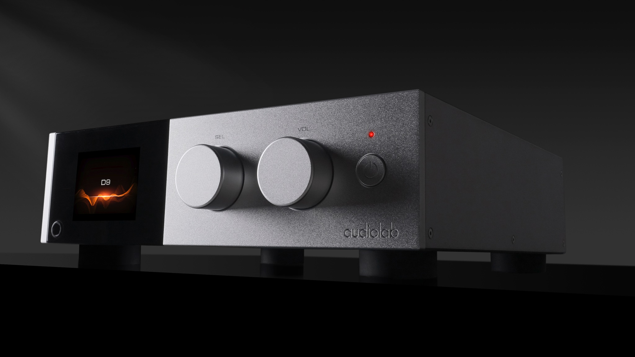 Audiolab D9 DAC. It is a silver rectangular hi-fi component with a dark display and a level meter in that display. The D9 is against a black background and on a black surface.