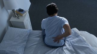 A man wearing a blue t-shirt sits up in bed rubbing his painful lower back
