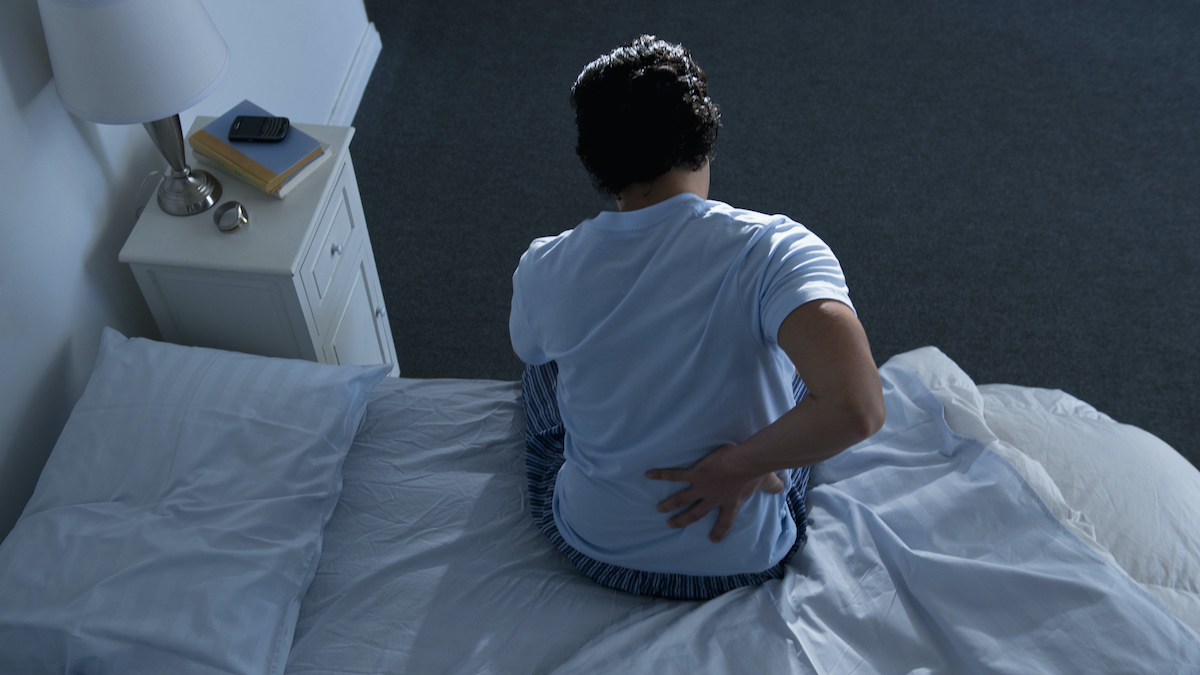 Seorang pria yang mengenakan kaos biru duduk di tempat tidur sambil menggosok punggung bawahnya yang sakit
