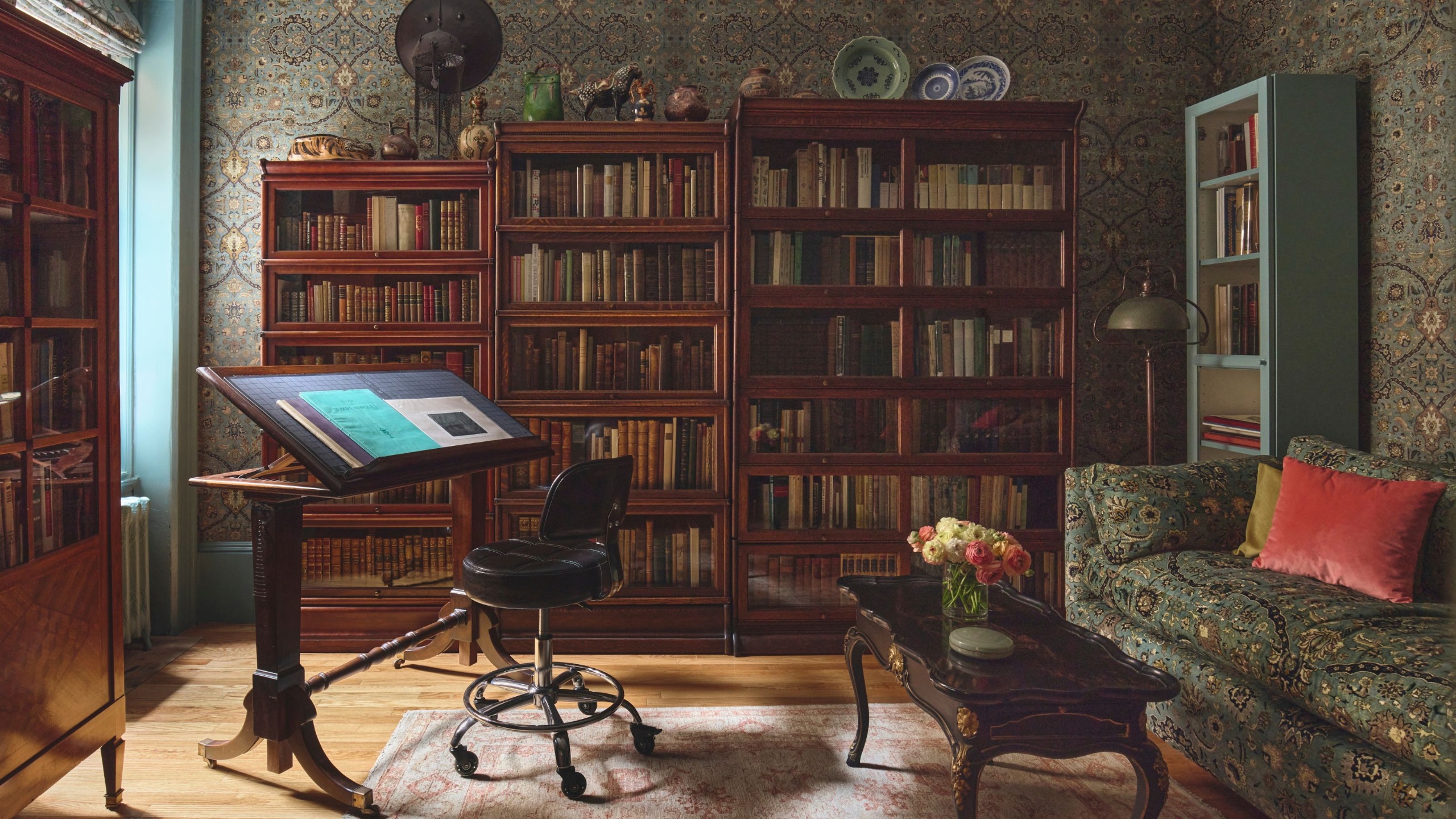 Cozy home library featuring dark wooden glass-door bookcases, a sloped reading desk with a rolling stool, patterned wallpaper, and a floral sofa.