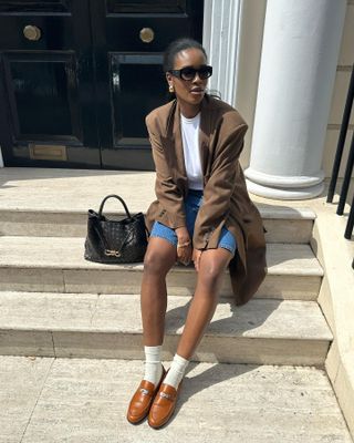 A woman wearing a brown blazer, white T-shirt, jean shorts, and brown loafers with a brown Bottega Veneta Andiamo bag.