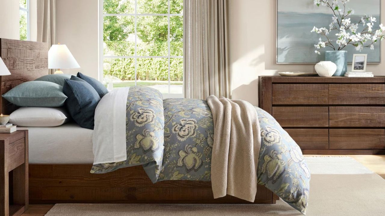 Side view of a dark wood bed frame dressed with blue and white bed linen; in the background, a vase of flowers on a dark wood dresser, and beige curtains covering windows looking onto a garden.