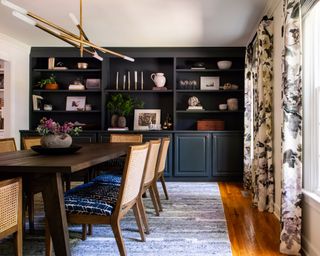 A dining room with wooden tale, rattan chairs, blue wall-to-wall shelving unit, black and white floral curtains and contemporary sputnik chandelier
