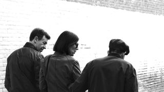 Three people walking, captured from the back, an old stonewall in the back in black and white color