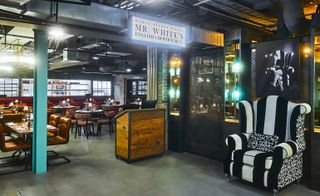 A restaurant in the New Road Hotel. A black and white upholstered armchair sits at the reception, with a wooden reception bar. Brown leather chairs, with both square and round tables are arranged throughout the area.
