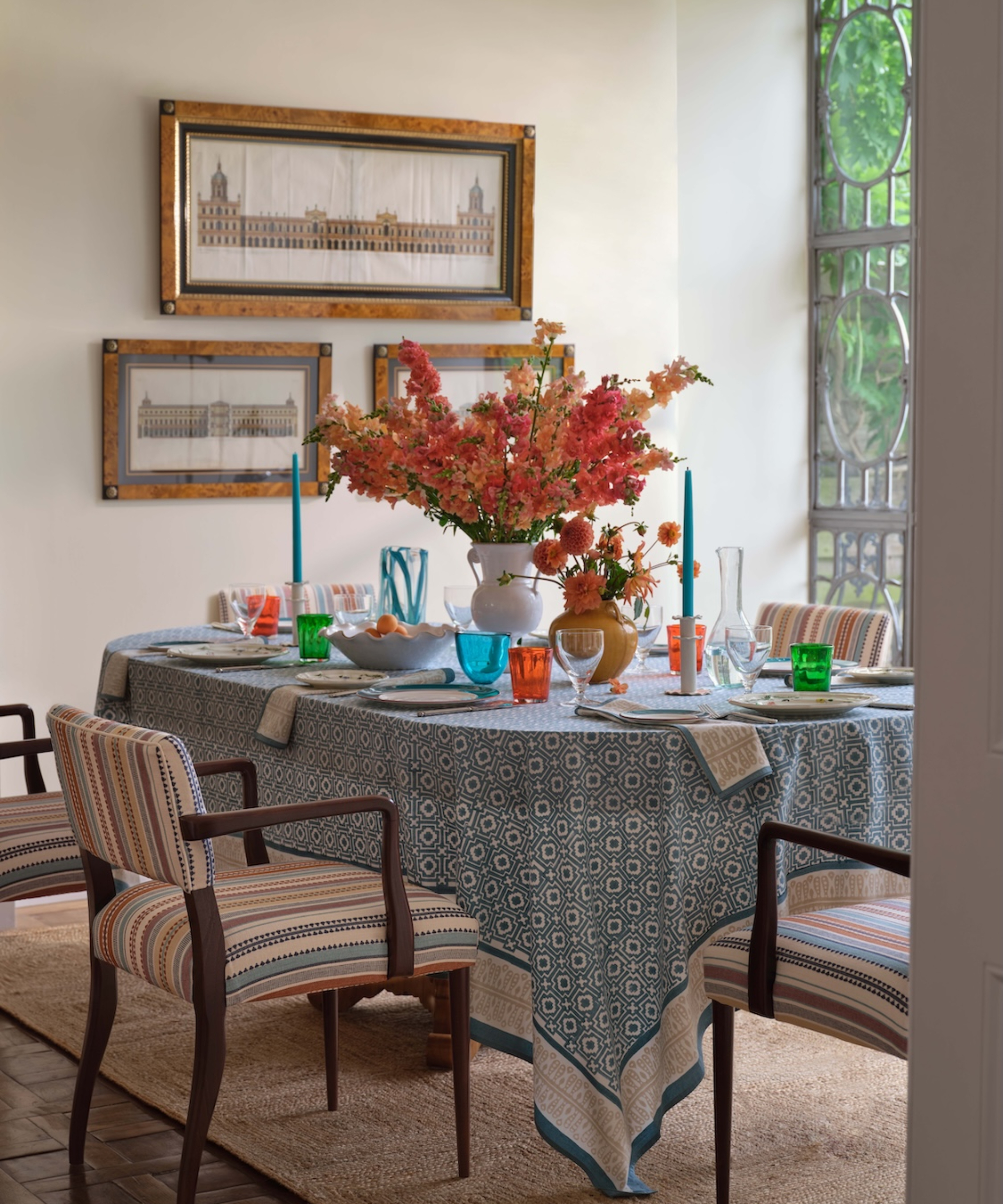 table covered in blue and white tablecloth, striped upholstered dining chairs, table set with candles and large floral arrangement