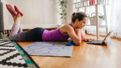 Woman lies on her front, head and hand propped up on a Pilates ball, looking at a tablet. Her hair is tied in a ponytail and she is wearing sportswear